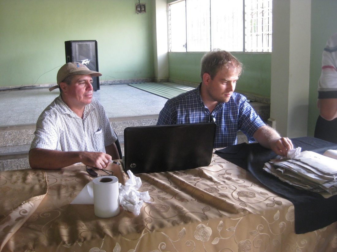 Don José Inez Gómez y Erlend M. Johnson sentados a una mesa con un portátil y un fajo de documentos sueltos sobre un paño